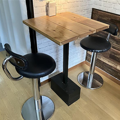 2名様用テーブル席は、デートや親しい友人との語らいにぴったりな、隠れ家のようなコンパクトな空間です。温かみのある木製天板のテーブルに、背もたれ付きのスタイリッシュな黒のハイチェアを設置。白レンガ調の壁が柔らかな雰囲気を醸し出し、二人だけの特別な時間を演出します。