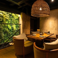 コンクリート打ちっぱなしの壁と、鮮やかな緑のウォールアートが調和する、洗練された空間。暖色の照明に照らされた天然木のテーブル席は、ゆったりと寛げる大人の隠れ家の雰囲気。わら編みのペンダントライトや温かみのある椅子が、モダンな中に自然の安らぎ。大切な人との語らいや、落ち着いた食事のひとときに最適