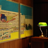 壁に飾られた大きなアート作品がおしゃれな店内を彩ります。人々が活動している様子や、建物の構造が細かく表現されており、賑やかな雰囲気を醸し出しています。また、横には緑色のデスクランプが置かれており、落ち着いた雰囲気をさらに引き立てています。全体的に、温かみのあるインテリアが感じられる空間です。