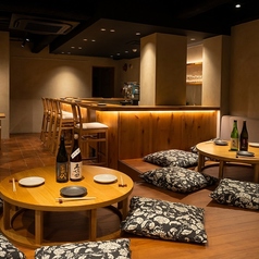 足を崩してゆったりと寛げる座敷エリアです。シックな黒い柄の座布団と低い丸座卓が、モダンでありながら和の安らぎを感じさせます。暖色系の照明が土壁風の壁を照らし、心地よい雰囲気を演出。親しい仲間と美味しい料理と美酒を囲み、心ゆくまで語り合える特別な空間です。