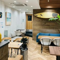 【リゾート気分を味わえる開放的な店内】白木とウッド調を基調とした明るい店内は、サーフボードやアートが飾られたおしゃれな雰囲気。奥にはゆったり寛げるソファ席、手前にはカジュアルに利用できるテーブル席が並び、シーンに合わせて選べます。居心地の良い空間で、リラックスしながら会話や食事を楽しめます。